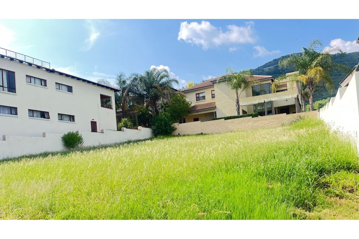 A view of a vacant residential plot featuring sloping terrain and surrounding boundary walls, with neighboring residential properties visible in the background.
