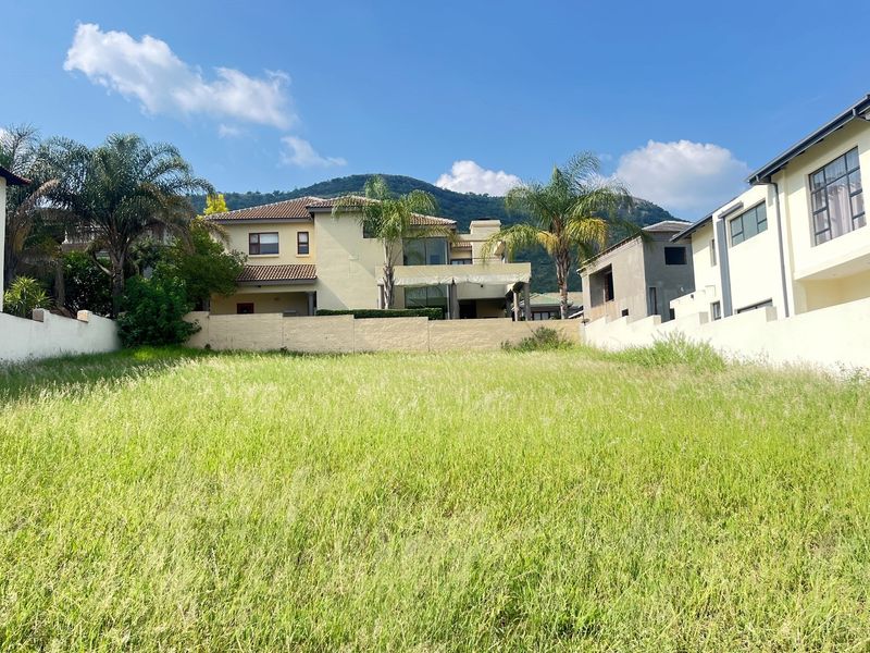 A vacant residential plot featuring tall grass, enclosed by perimeter boundary walls, with established neighboring properties and mountain views in the background.
