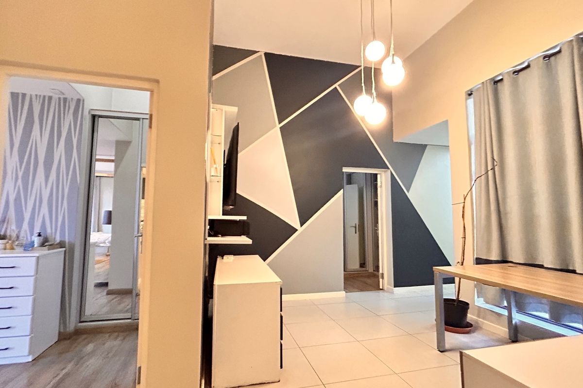 A bright living area featuring tiled flooring, modern pendant lighting, and a geometric accent wall, providing access to adjacent rooms.