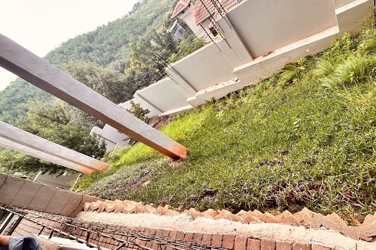 A rear garden area featuring a sloped lawn, boundary wall, and structural concrete pillars showing signs of unfinished construction and weathering.
