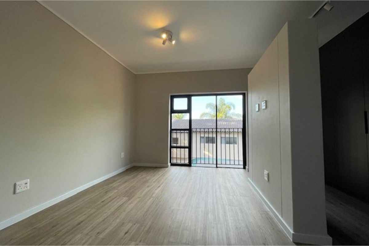 A bright, empty bedroom featuring modern laminate flooring, neutral walls, and a sliding glass door with a security gate leading to an outdoor area.