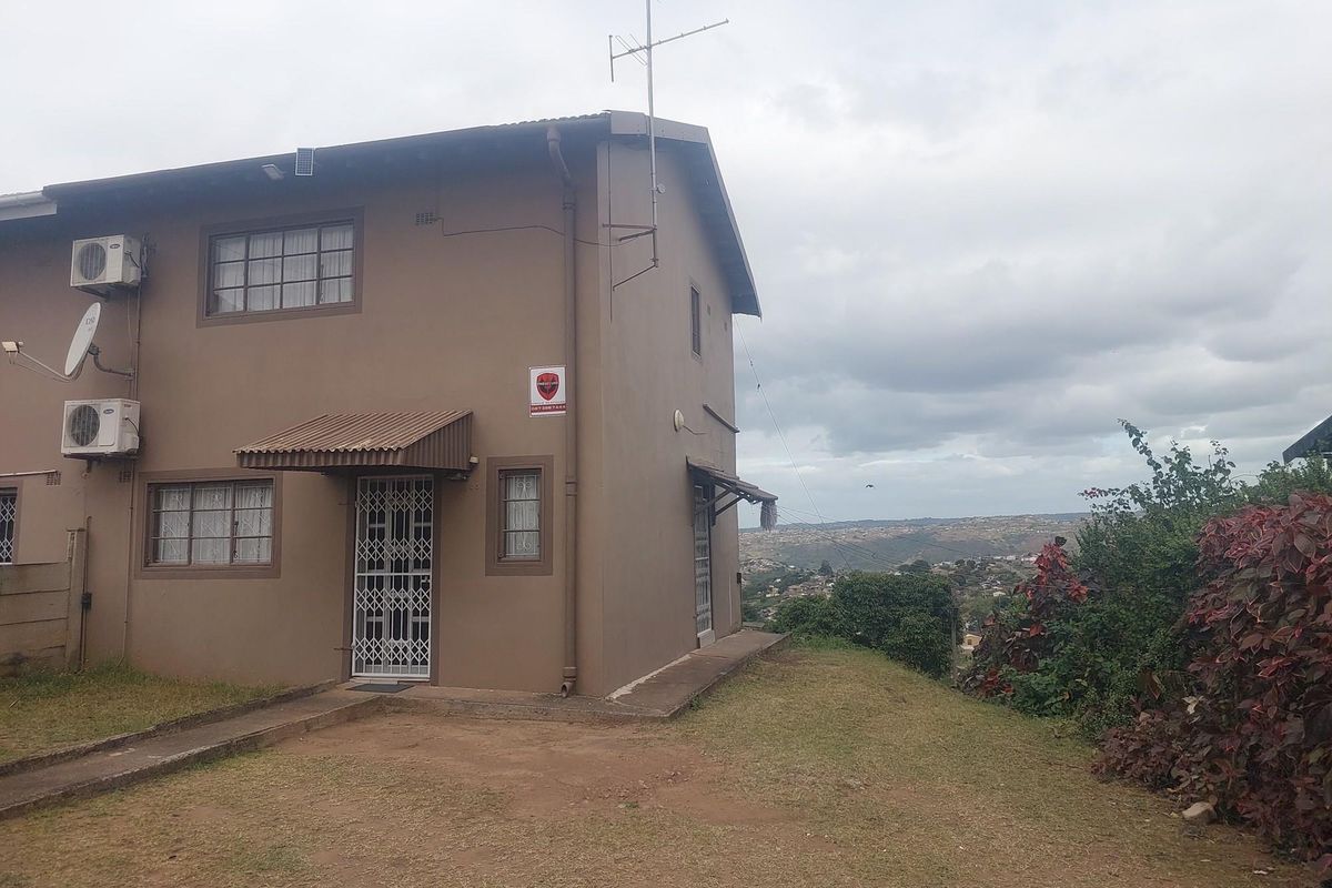 Exterior view of a two-story residential building featuring security gates, air conditioning units, and a sloped garden area with distant landscape views.