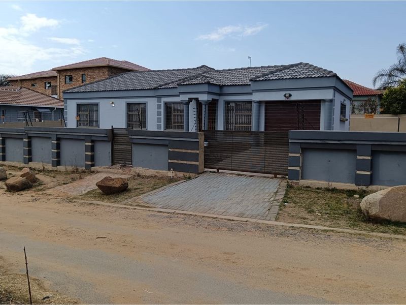 Front exterior view of a single-story house featuring a grey facade, tiled roof, secure perimeter wall, and a gated driveway entrance.