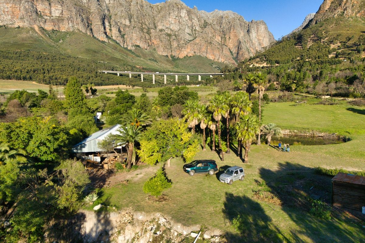 Aerial view of a farm property featuring a rustic outbuilding, mature palm trees, and expansive natural grounds set against a dramatic mountain backdrop.