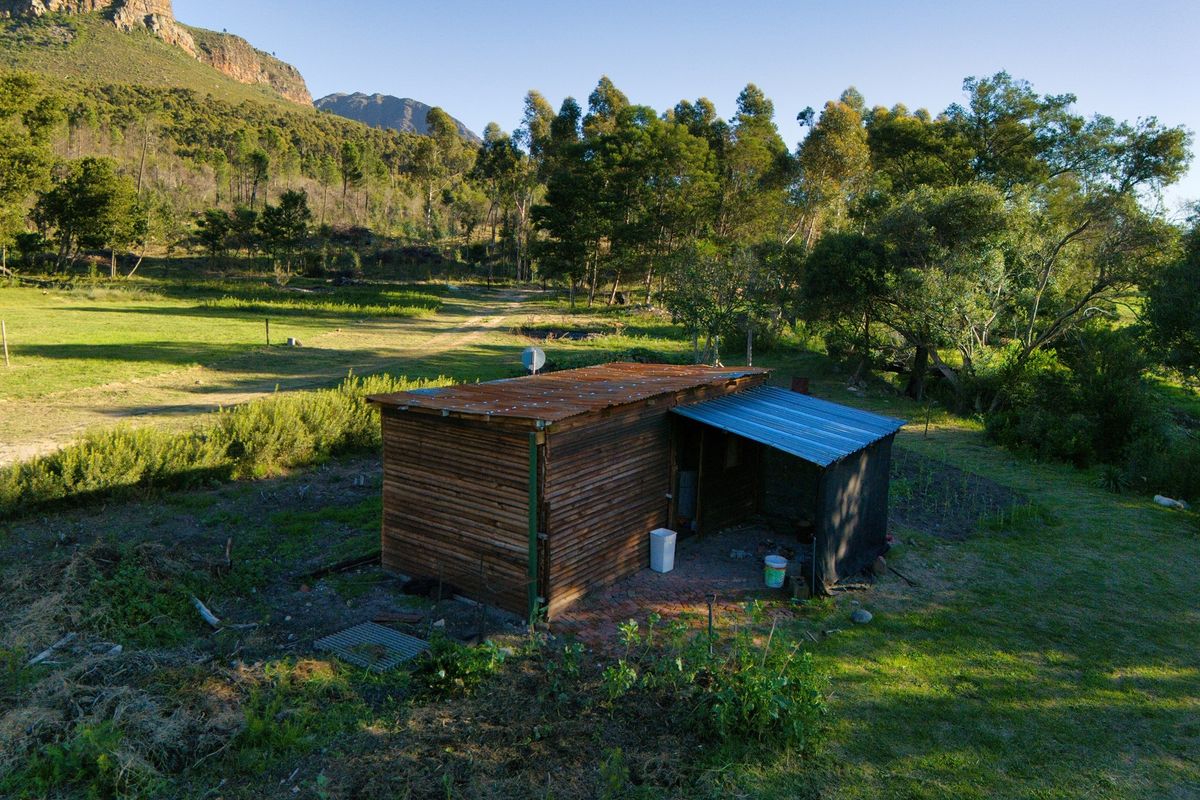 An aerial view of a rustic timber-clad structure with a corrugated metal roof, situated in a rural farm setting with surrounding trees and mountain views.