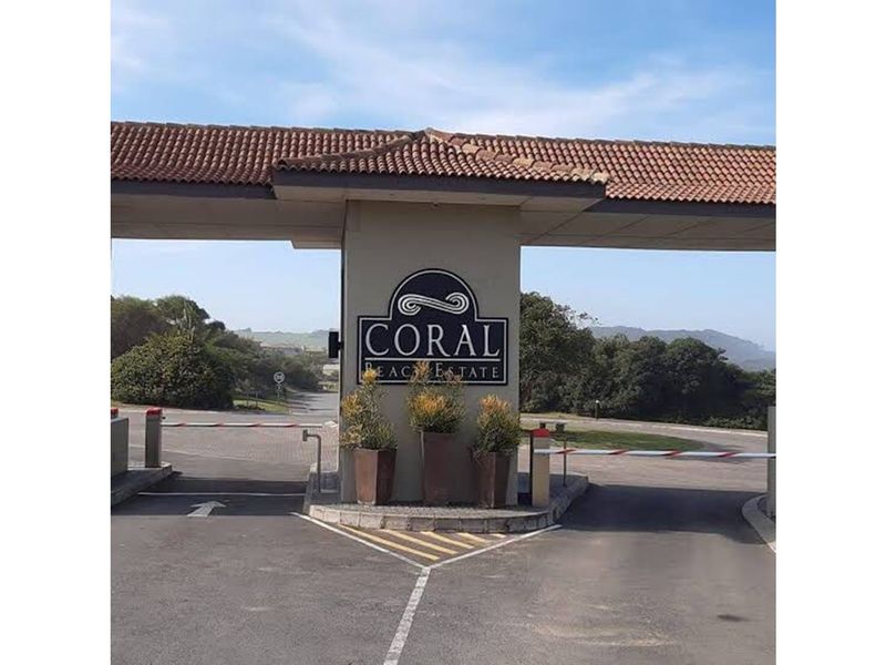 The entrance gate to Coral Beach Estate, featuring a paved driveway, security boom gates, and estate signage under a tiled roof structure.