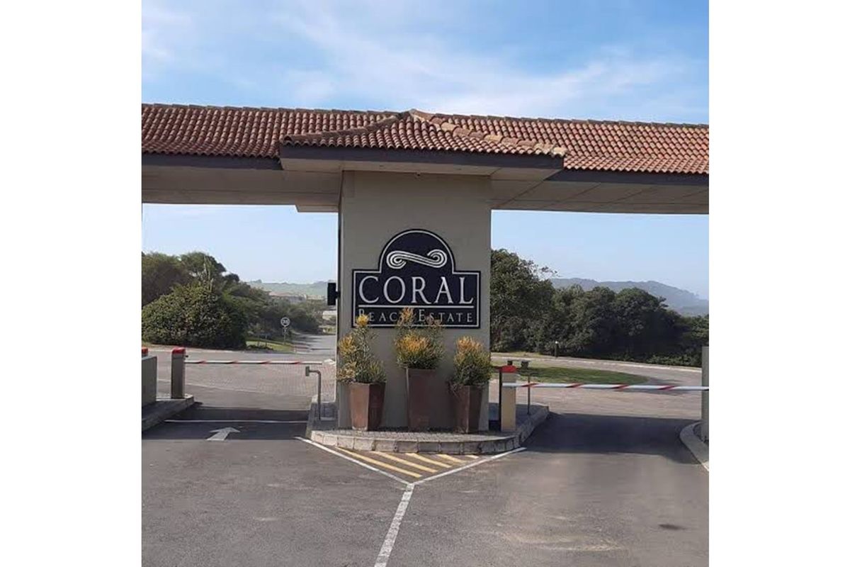 The entrance gate to Coral Beach Estate, featuring a paved driveway, security boom gates, and estate signage under a tiled roof structure.