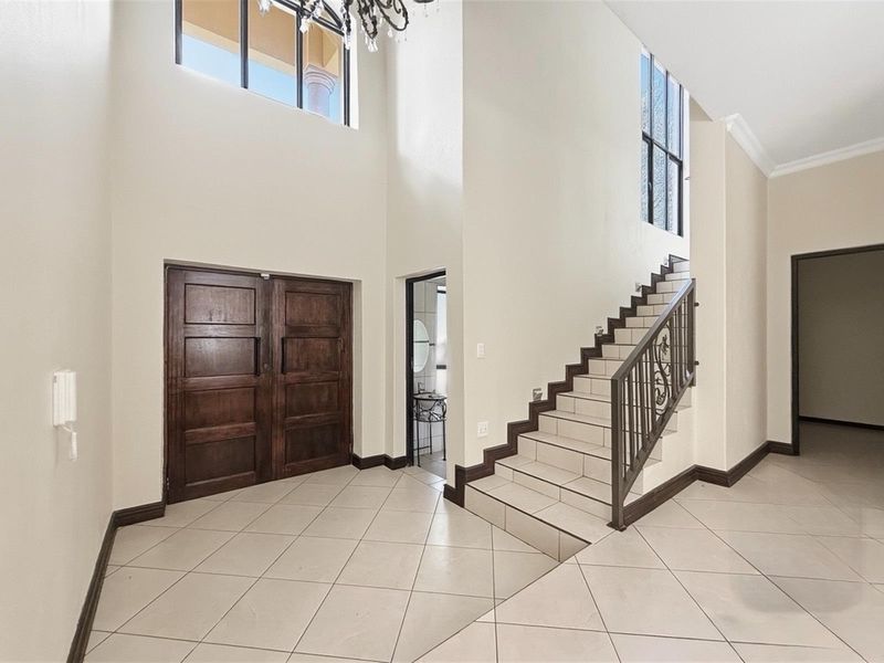 Spacious double-volume entrance hall featuring tiled flooring, a grand staircase with wrought iron balustrade, and high windows allowing for abundant natural light.