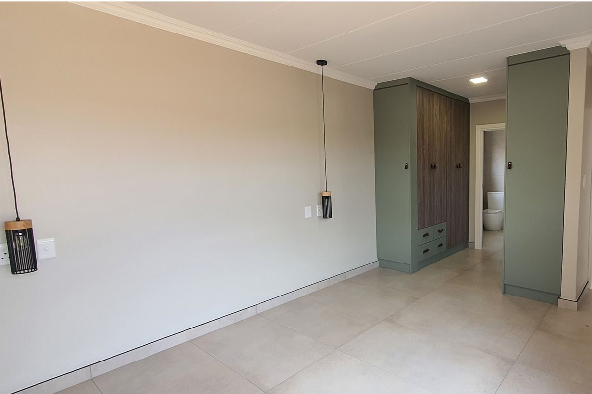 A modern bedroom featuring built-in sage green cabinetry with wood-grain accents, tiled flooring, and contemporary pendant lighting, leading to an en-suite bathroom.