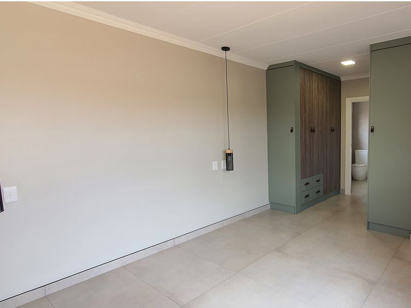 A modern bedroom featuring built-in sage green cabinetry with wood-grain accents, tiled flooring, and contemporary pendant lighting, leading to an en-suite bathroom.