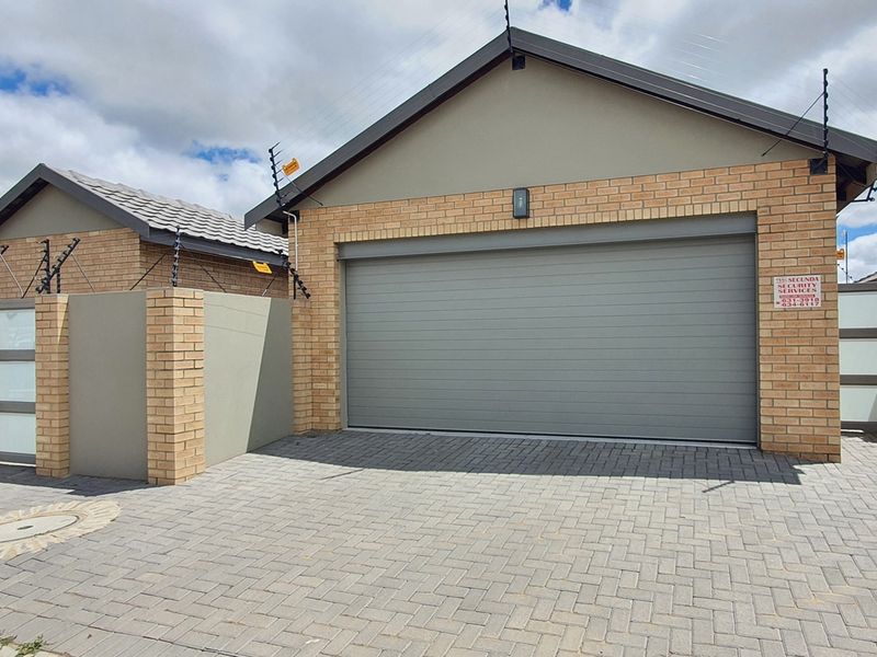 A modern residential garage facade featuring a paved driveway, brick and plaster exterior, and secure perimeter fencing with electric security wires.