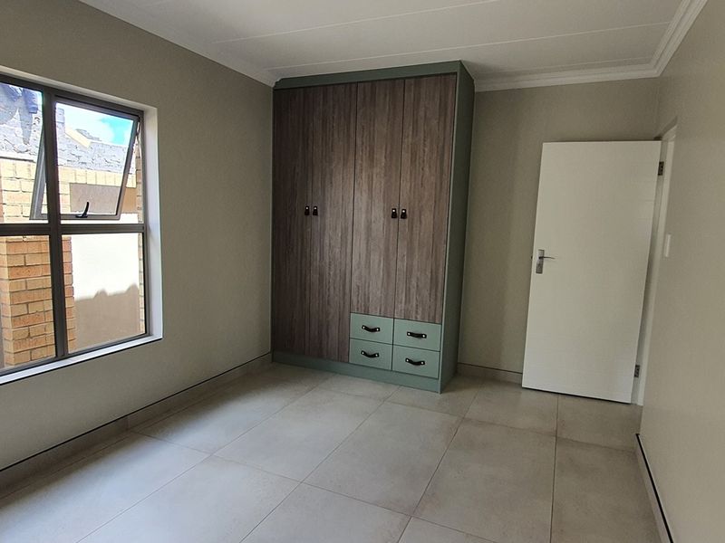 A clean, modern bedroom featuring neutral floor tiles, a built-in wardrobe with wood-grain and sage green accents, and a large window.