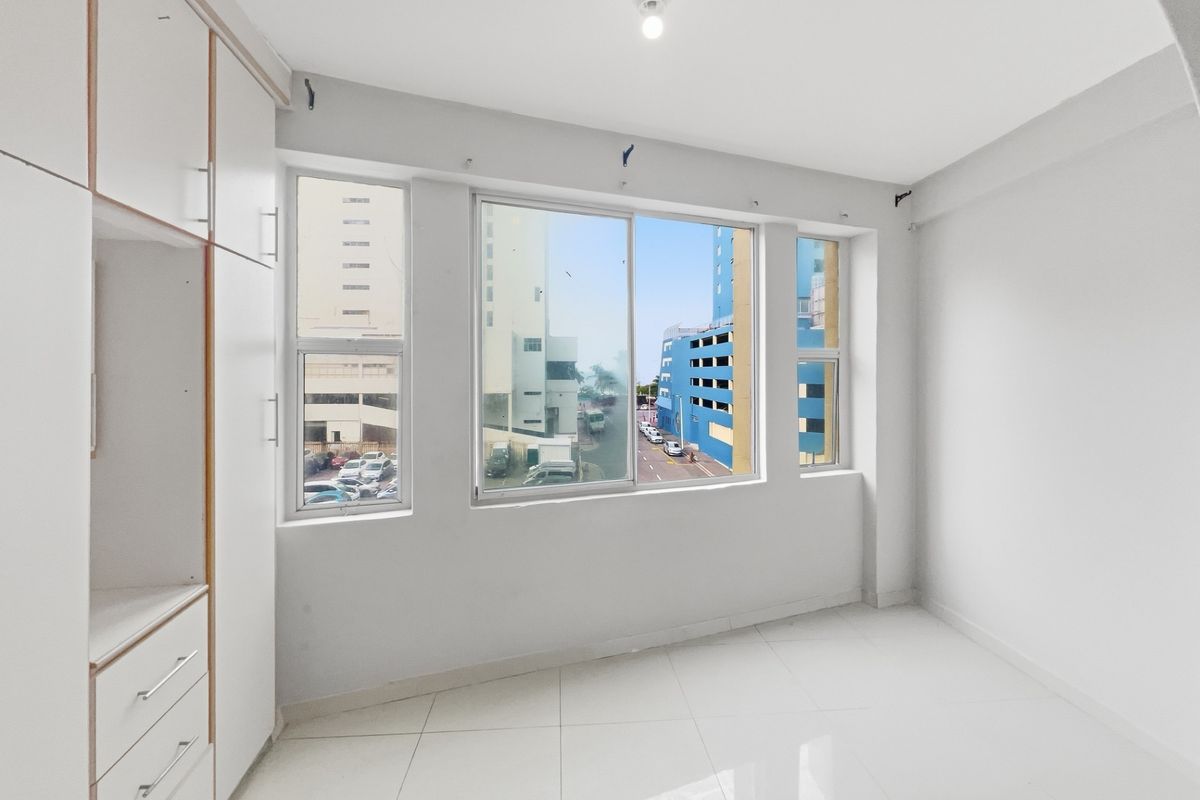 An empty bedroom featuring white tiled flooring, built-in cupboards, and large windows offering an urban street view.