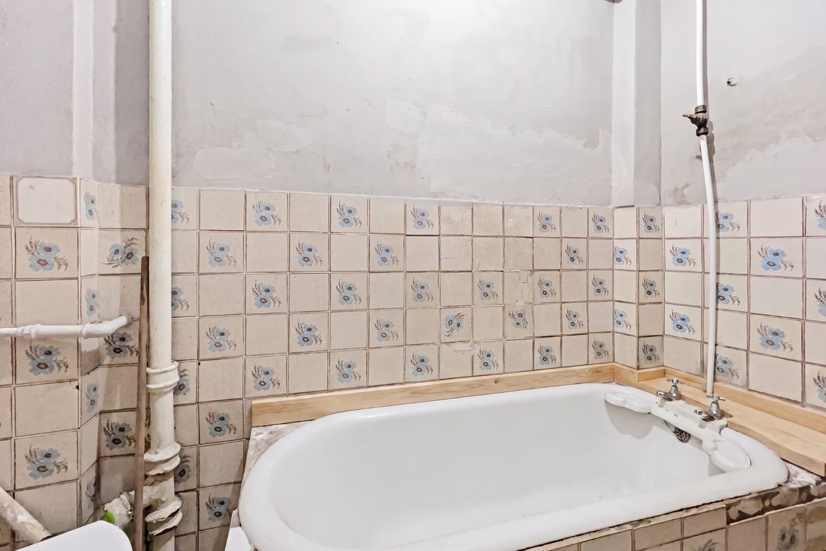 A dated bathroom featuring a bathtub with floral tiled splashback and exposed plumbing, requiring significant renovation and cosmetic updates to improve its condition.