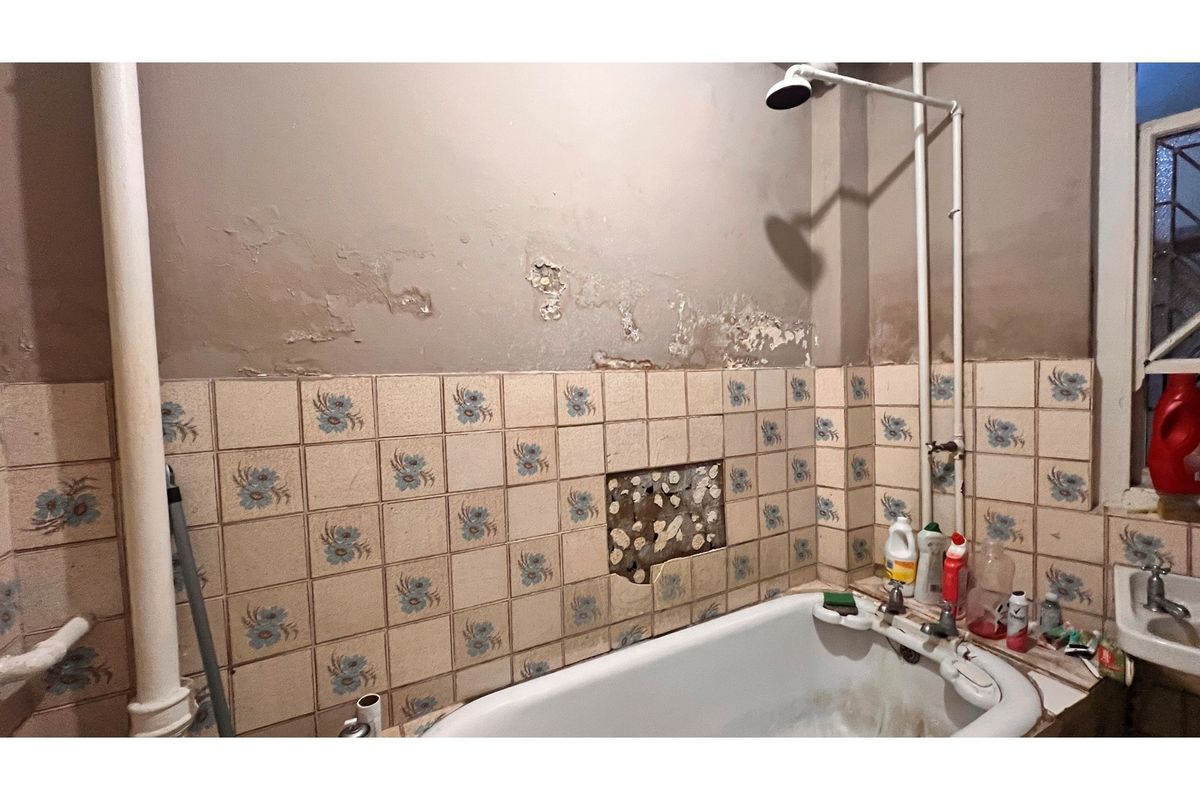 A bathroom in need of renovation, featuring a bathtub, exposed plumbing, and significant damage to the wall tiles and plaster finishes.