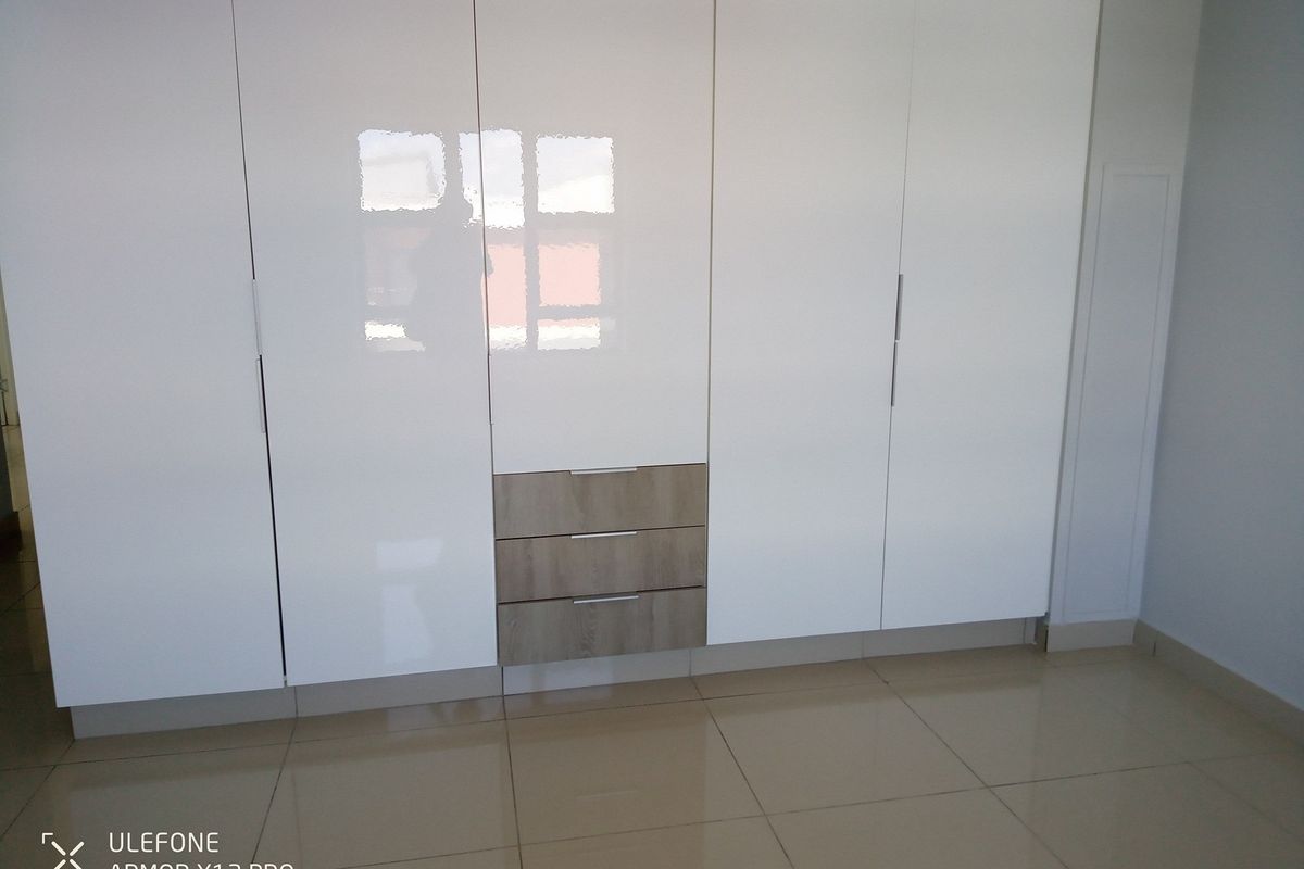 A modern bedroom featuring floor-to-ceiling built-in wardrobes with high-gloss white doors and integrated timber-look drawers, complemented by neutral tiled flooring.