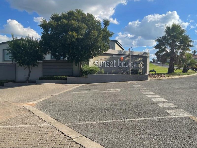 Entrance view of the Sunset Boulevard residential complex featuring paved driveway, perimeter wall, and signage.