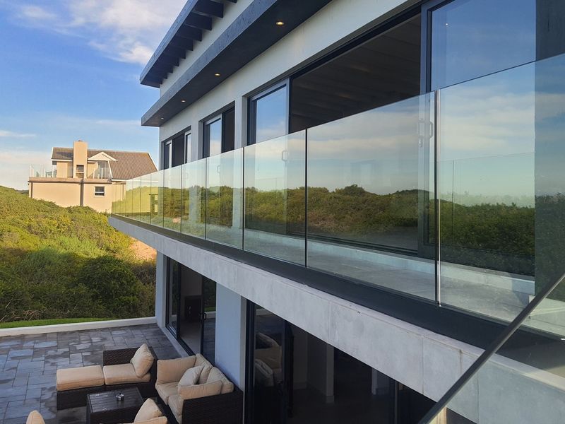 Modern upper-level balcony featuring a frameless glass balustrade, recessed lighting, and large sliding doors overlooking a landscaped coastal environment.