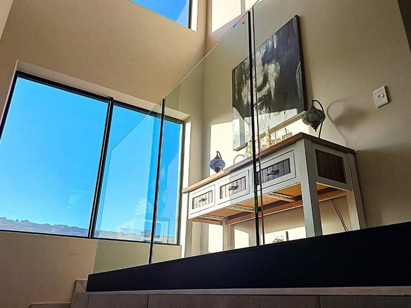 A bright, modern hallway featuring a sleek glass balustrade, double volume ceilings, and expansive windows that provide abundant natural light.