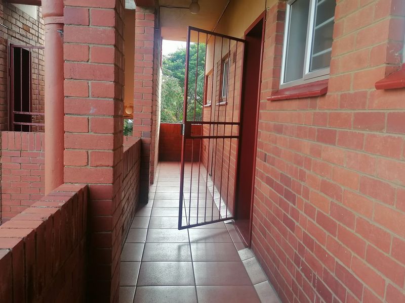 An exterior apartment walkway featuring exposed brick walls, tiled flooring, and a metal security gate providing access to the unit.