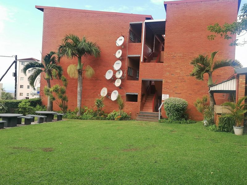 Exterior view of a brick apartment building featuring a communal lawn area, concrete seating, and external stairwell access.