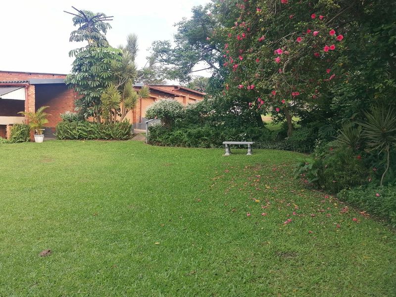 A well-maintained garden area featuring a lush green lawn, mature trees, and a view of the brick building exterior and covered patio.