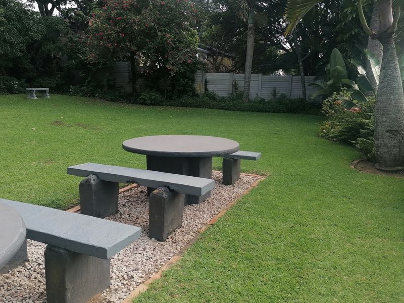A well-maintained garden area featuring established lawn, mature trees, perimeter boundary wall, and fixed concrete seating arrangements.