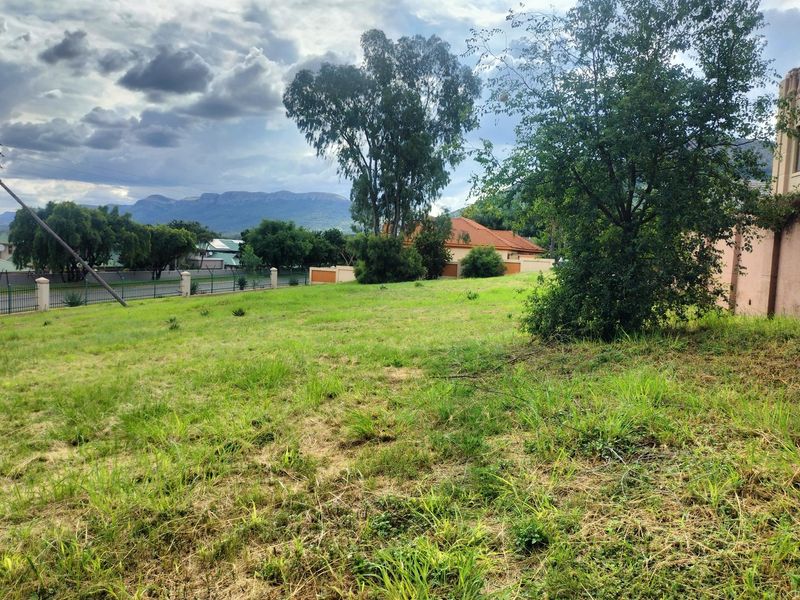 A vacant residential plot featuring established trees, perimeter fencing, and scenic mountain views in a quiet neighborhood setting.