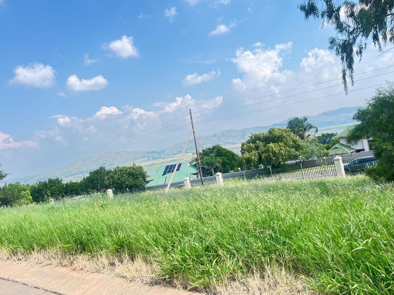 A view of a vacant residential plot featuring tall grass, perimeter fencing, and distant mountain views under a clear blue sky.