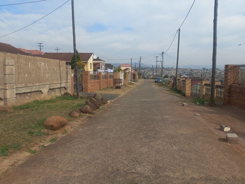 A street view showing the paved road, surrounding residential boundary walls, and overhead utility infrastructure in a suburban neighborhood setting.