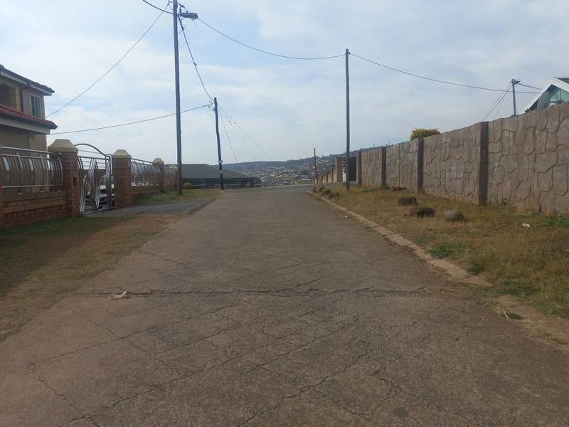 A view of the residential street approach featuring asphalt road surface, perimeter boundary walls, and established utility infrastructure.