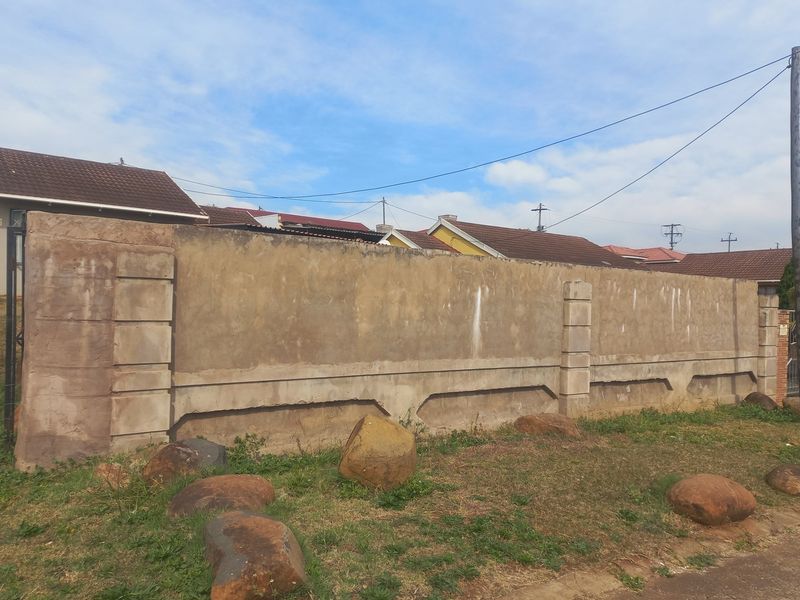 A street-facing view of a weathered precast concrete boundary wall with integrated pillars and a metal gate entrance.