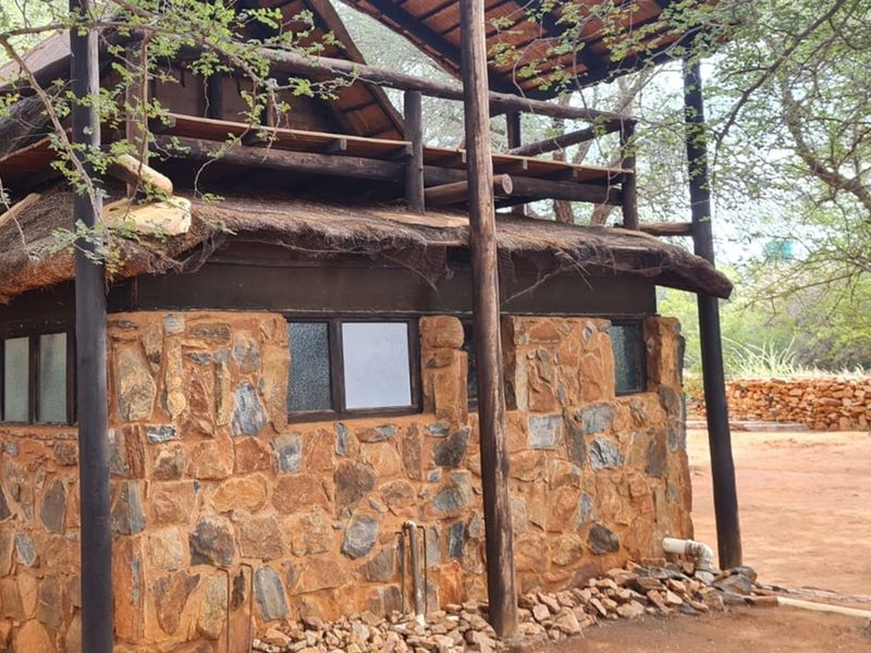 Rustic stone-clad farm building featuring a thatched roof and an elevated wooden deck structure, set within a natural bushveld environment.