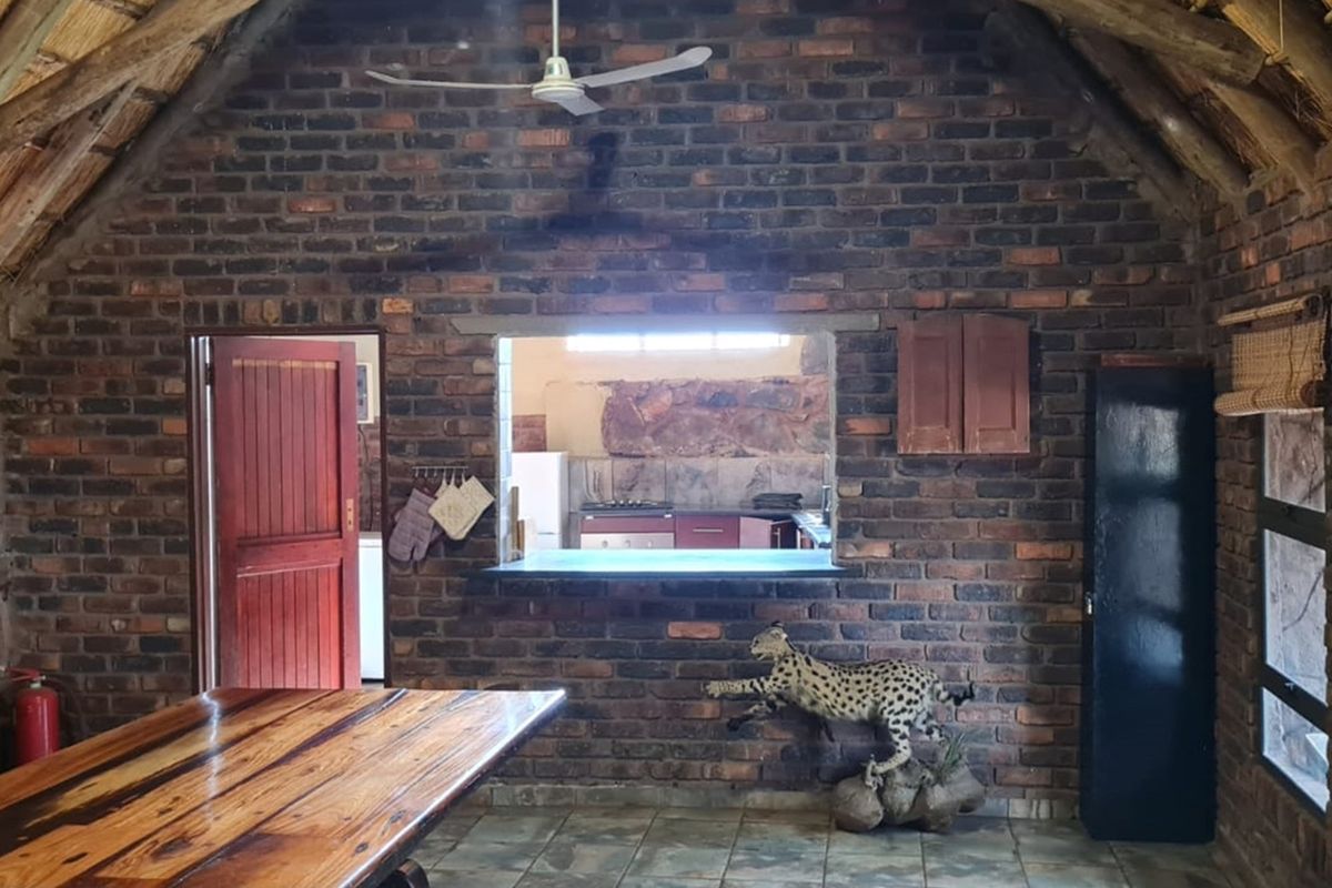 Rustic living area featuring exposed brick walls, a high thatched roof, ceiling fan, and a service window looking into the kitchen.