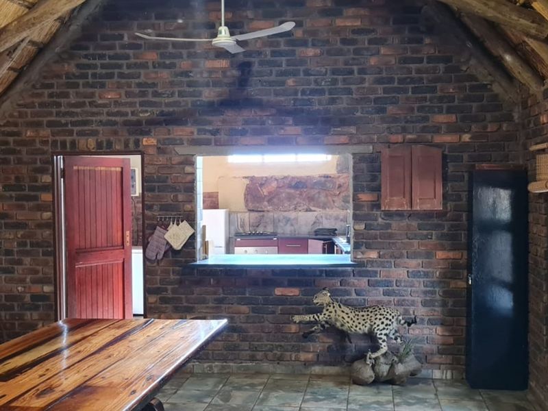 Rustic living area featuring exposed brick walls, a high thatched roof, ceiling fan, and a service window looking into the kitchen.