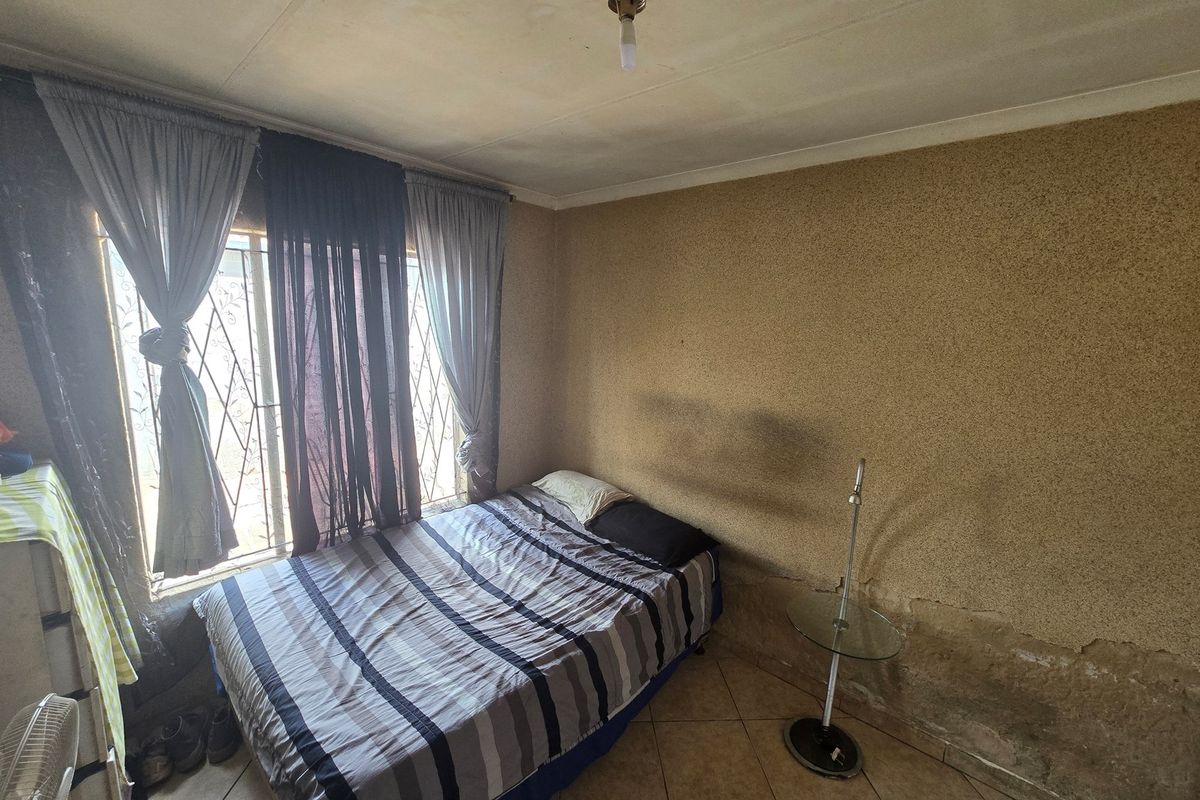 A bedroom featuring tiled flooring and a window with security bars, currently showing significant wear to the wall finishes.