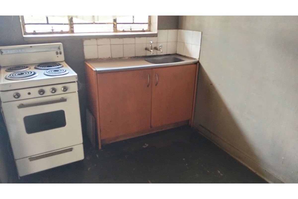 A basic, dated kitchen area featuring a freestanding stove, single sink, and simple base cabinetry with unfinished wall surfaces.