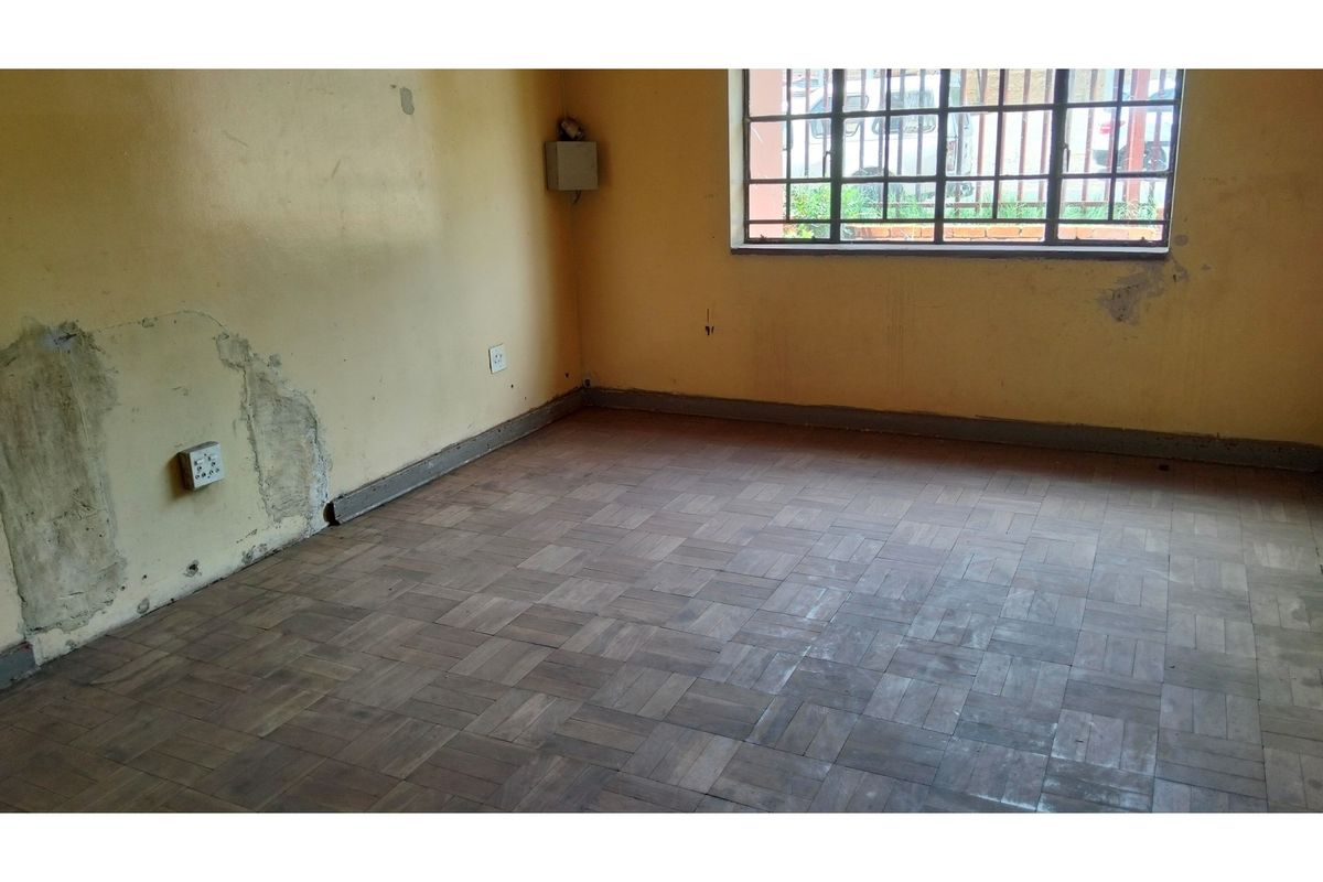 An empty living room featuring parquet flooring, security bars on the window, and walls requiring plaster repair and repainting.