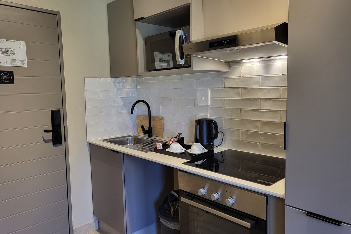 Compact modern kitchenette featuring grey cabinetry, subway tile backsplash, built-in oven, electric hob, extractor fan, and a sleek black faucet.