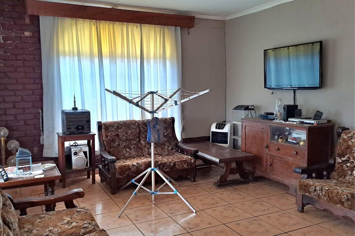 A living room featuring tiled flooring, a brick accent wall, and large window with curtains, offering a dated but functional living space.