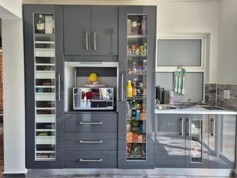 A modern scullery featuring dark grey cabinetry, integrated microwave, glass-fronted pantry storage, and a compact stainless steel sink with tiled splashback.