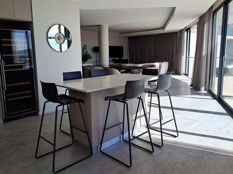 Modern open-plan living area featuring a central kitchen island, built-in wine cooler, tiled flooring, and expansive sliding glass doors leading to a balcony.