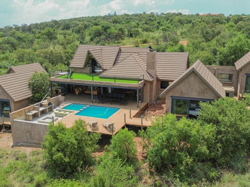 Aerial view of a modern residence featuring a swimming pool, expansive timber decking, built-in braai area, and a balcony set within a natural landscape.