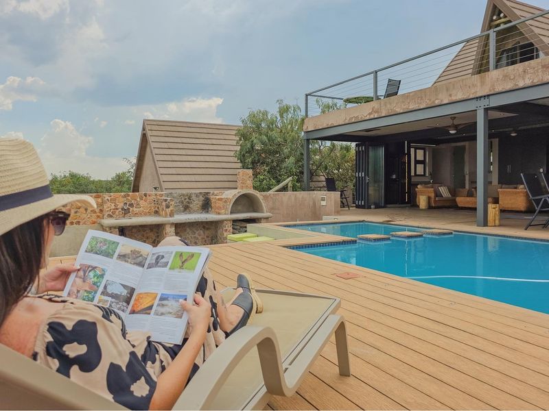 Outdoor pool area featuring timber decking, a built-in stone braai, and a covered patio space overlooking the swimming pool.