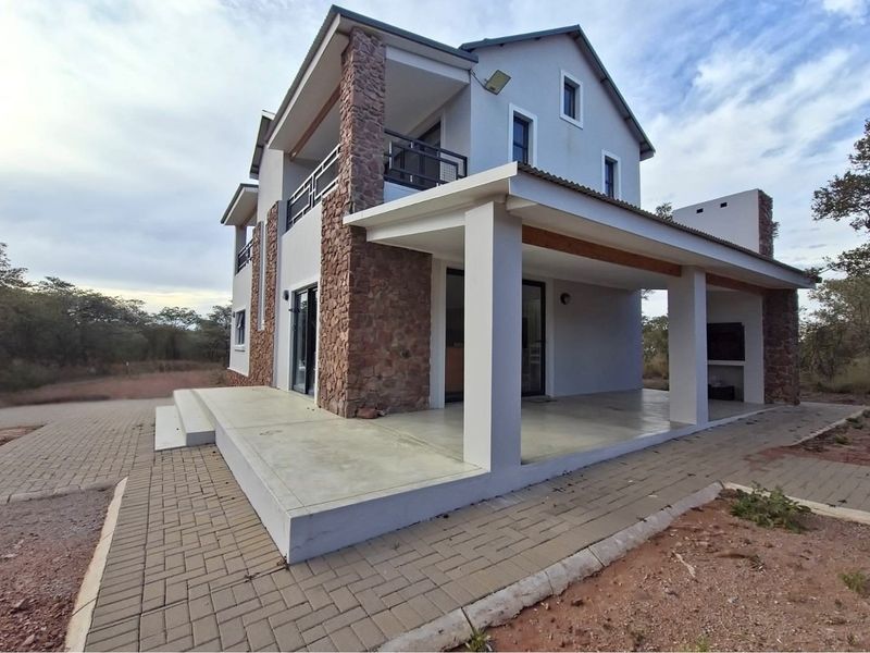 A modern double-story house featuring stone cladding accents, a spacious covered patio, and a paved driveway in a natural bushveld setting.