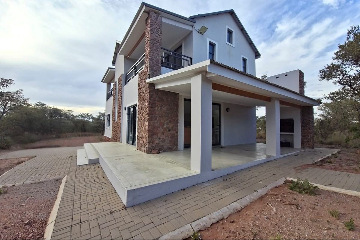 A modern double-story house featuring stone cladding accents, a spacious covered patio, and a paved driveway in a natural bushveld setting.