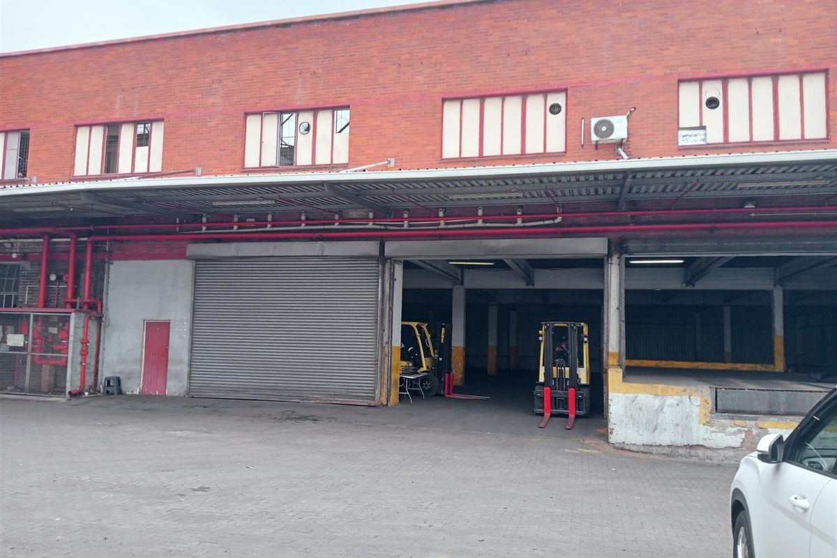 Industrial warehouse exterior featuring a large loading bay with roller shutter doors, covered canopy, and exposed fire suppression piping.
