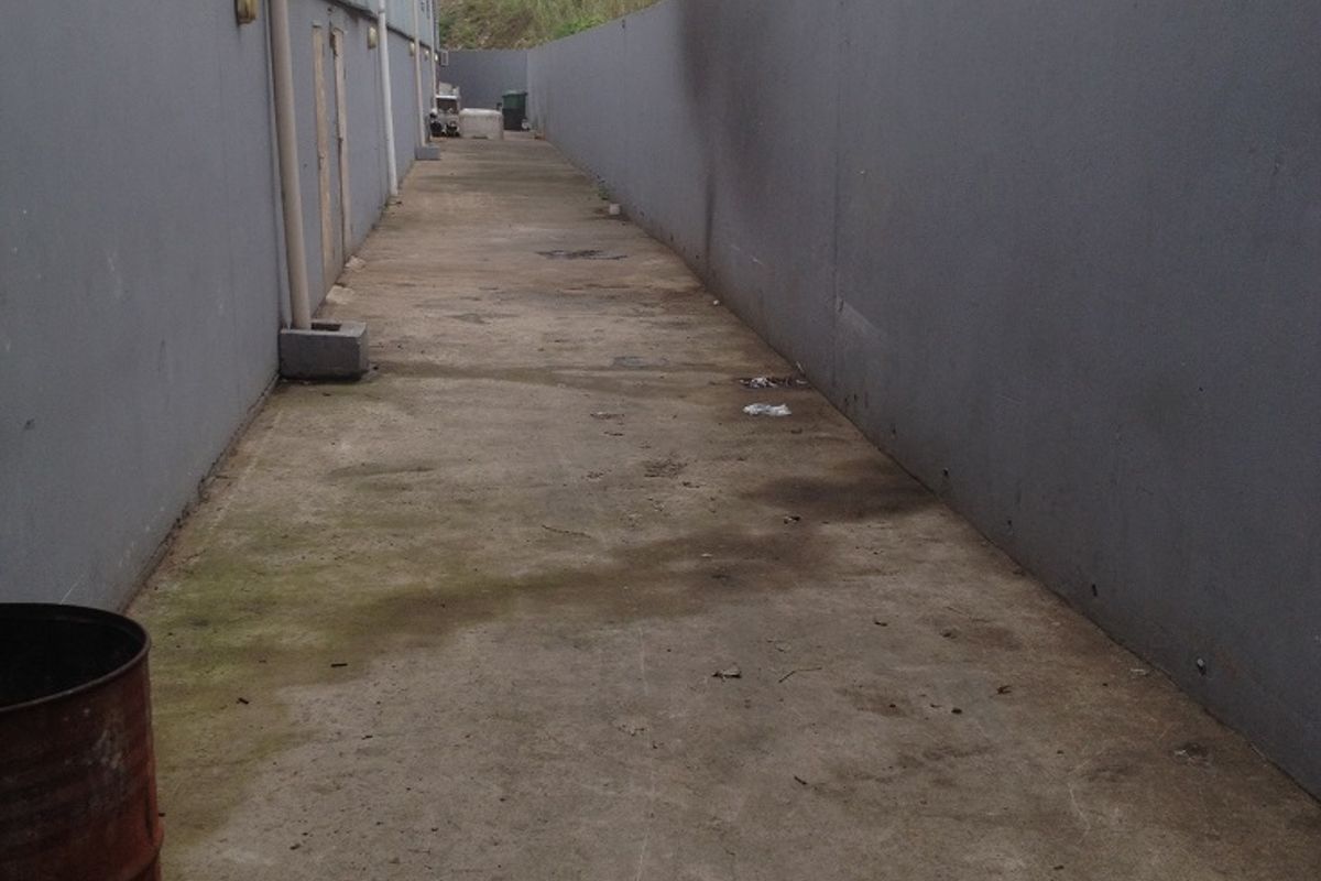 A narrow exterior side passage or service alley featuring concrete flooring and high boundary walls, suitable for industrial utility access.