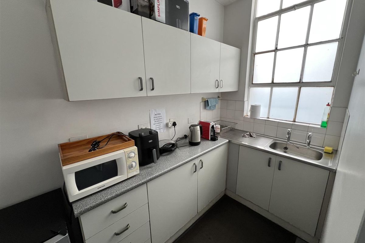 A compact industrial-style kitchenette featuring white cabinetry, a stainless steel sink, laminate countertops, and a large multi-pane window providing natural light.