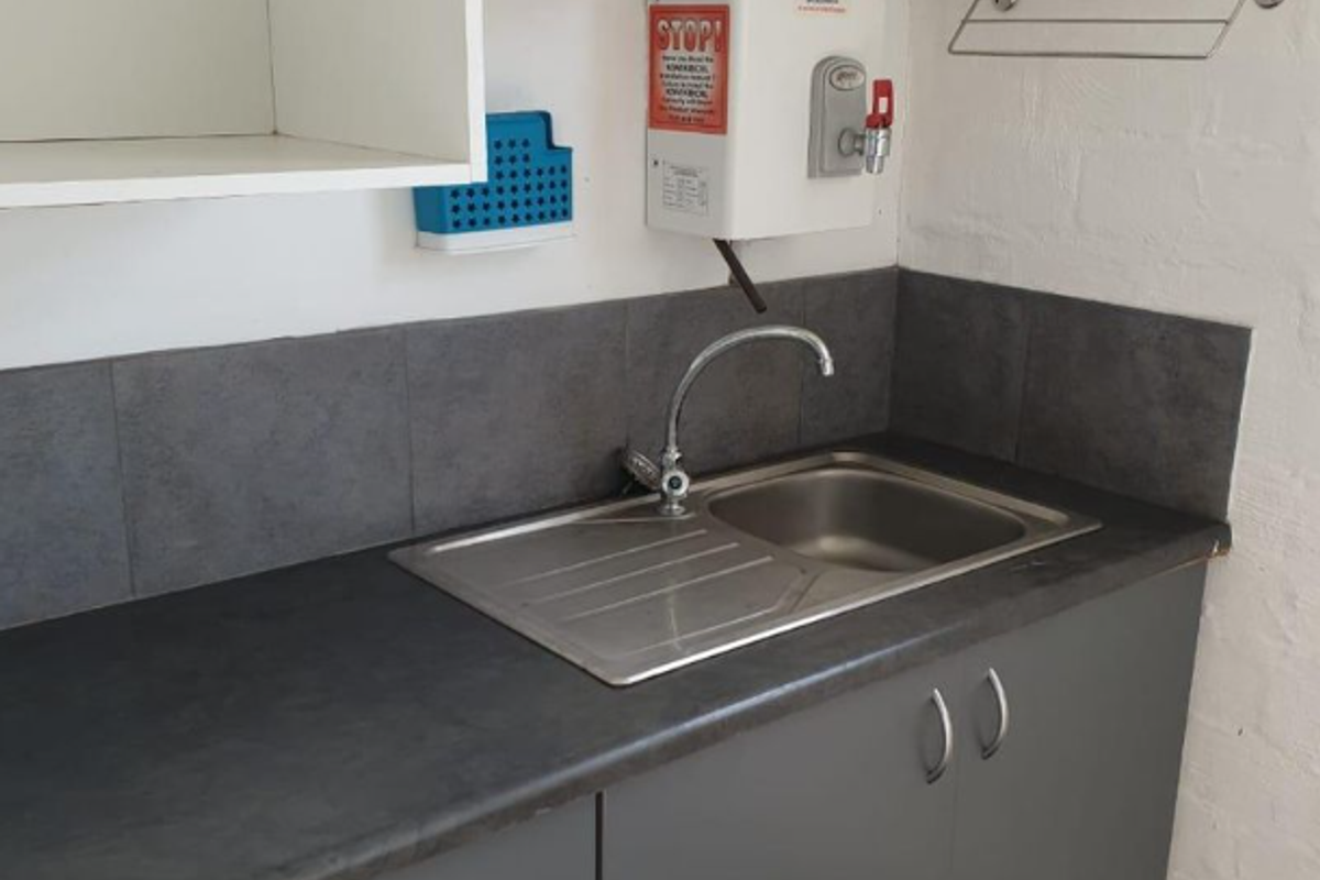 A functional kitchenette area featuring dark grey cabinetry, a stainless steel sink, tiled splashback, and a wall-mounted water heater.